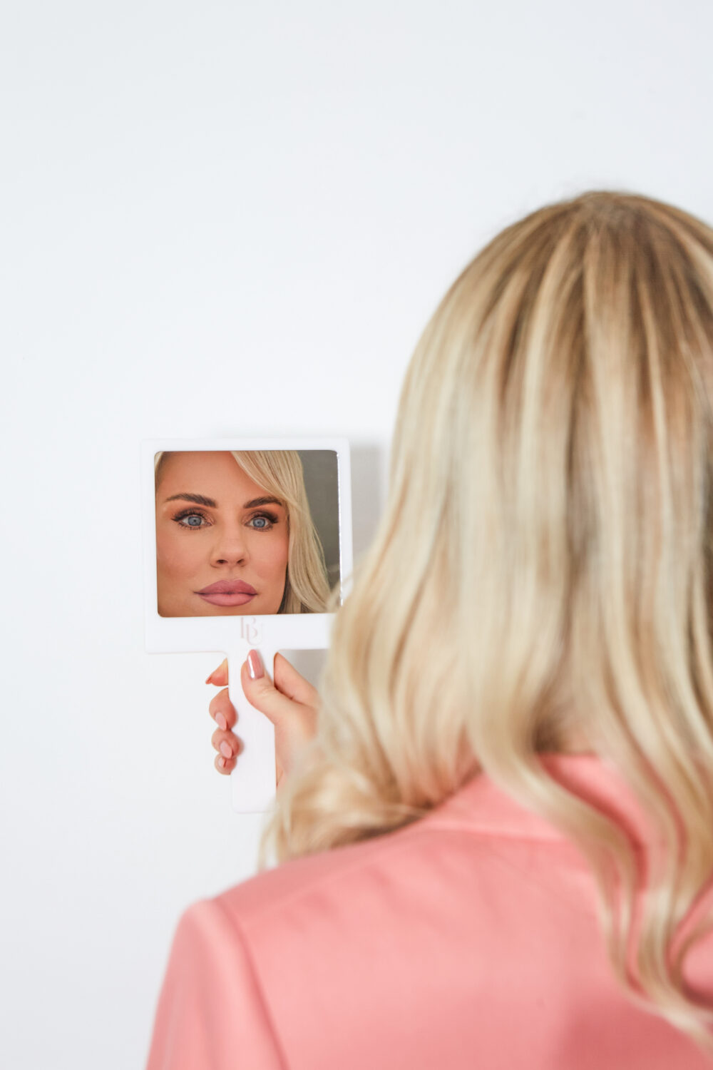 A photo of Caroline from BU Aesthetics wearing a salmon jacket looking at herself in a mirror